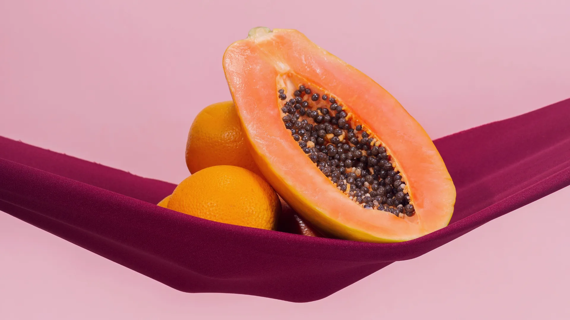 Aufgeschnittene Papaya und Orangen auf beerenrotem Stoff, warm ausgeleuchtet.